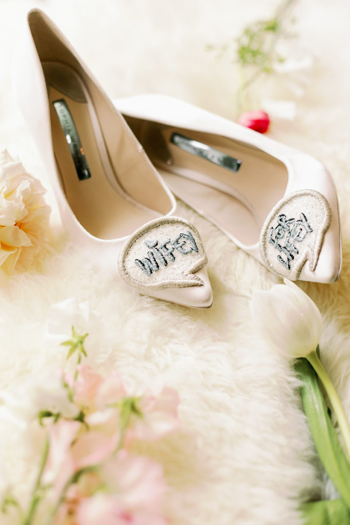 The bride's white pumps displayed on a textured white backdrop. The shoes have a patch on the toe that reads "wifey for lifey."