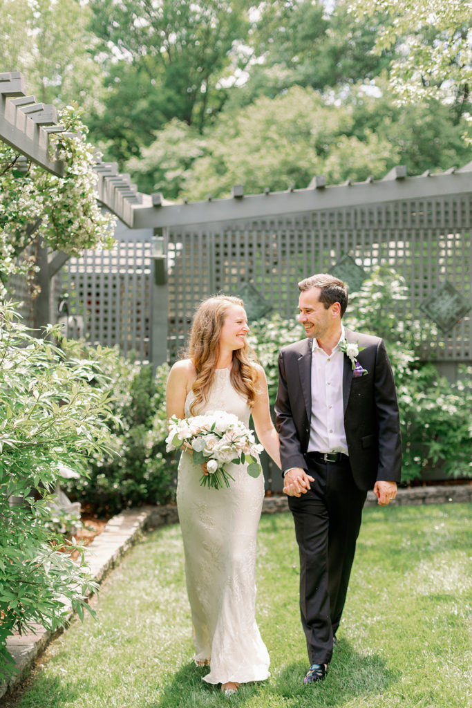 The bride and groom pose in their backyard in front of a grey painted fence adorned with climbing flowers.