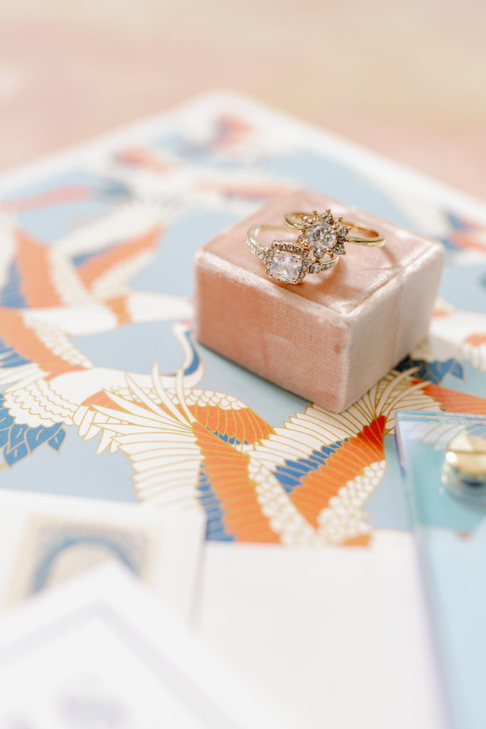 Stunning art-deco style rings are perched atop a muted pink velvet block. This block is displayed on top of pieces of the wedding invitation suite, including a brightly colored envelope liner. 